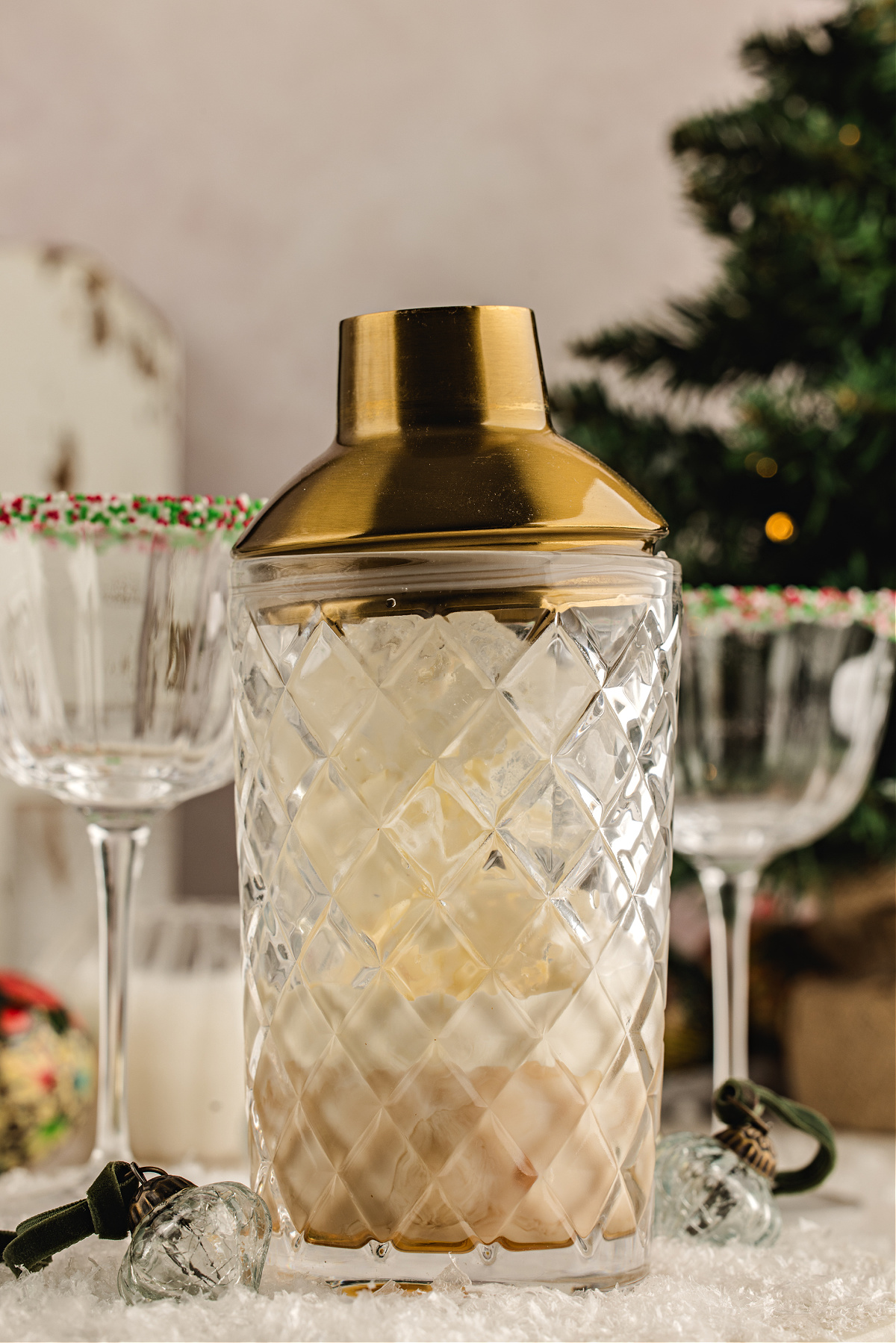 Cream liqueur poured over ice in a mixing glass to make a festive holiday cocktail. Irish cream being poured from a gold jigger into a mixing glass filled with ice and amaretto, with sprinkle-rimmed holiday coupes in the background.