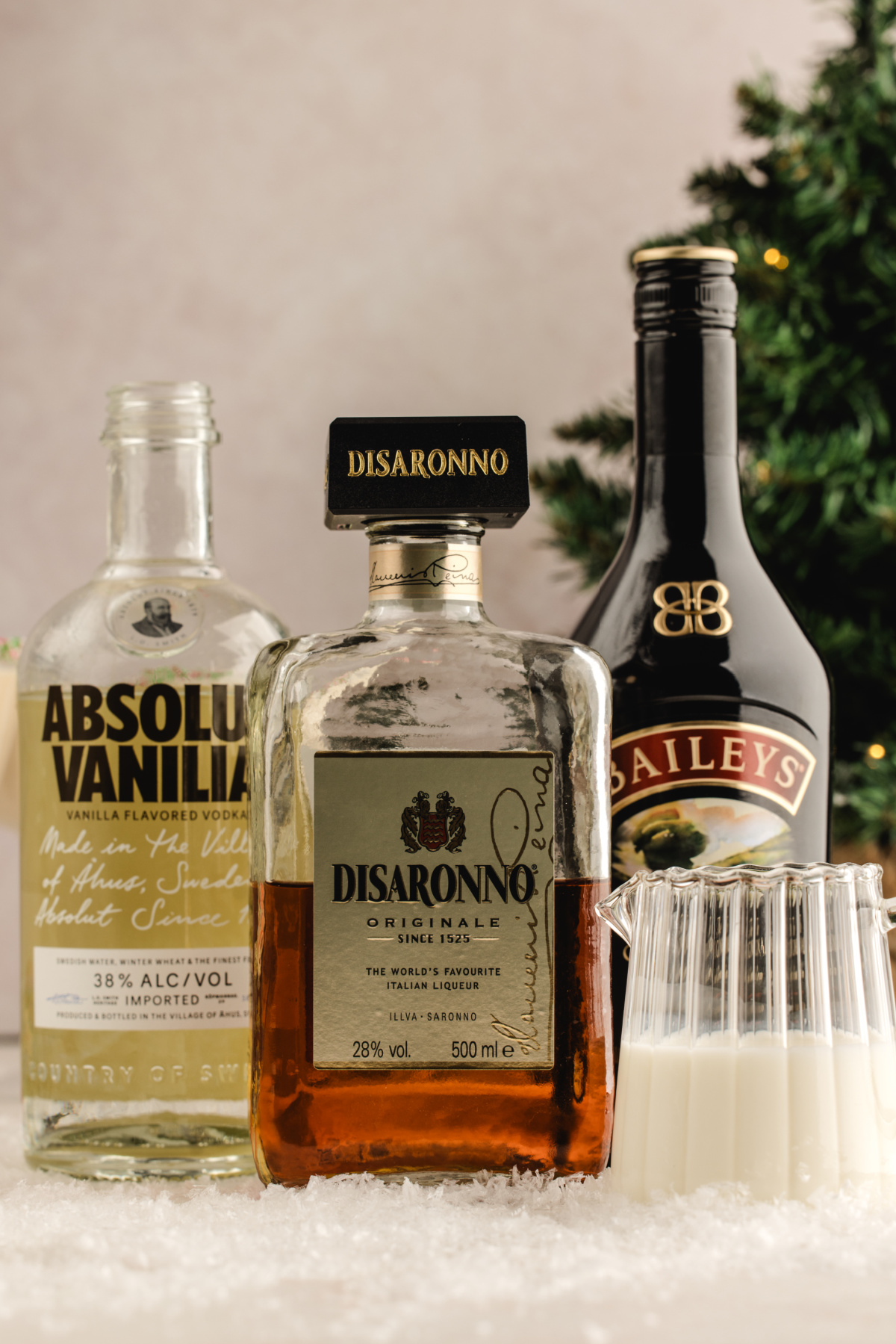 Holiday martini ingredients including vanilla vodka, amaretto, Irish cream, and cream on a snowy tabletop. Vanilla vodka, amaretto, Irish cream, and a small pitcher of cream arranged together on a snowy surface with a Christmas tree blurred behind them.