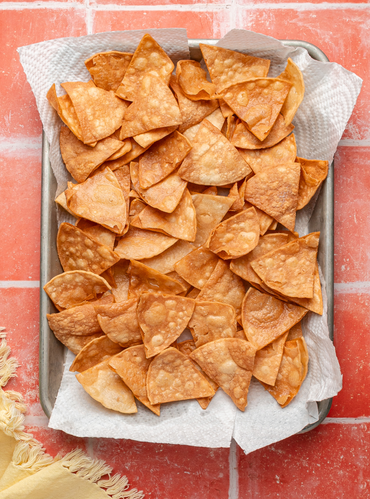 Freshly fried tortilla chips cooling on a sheet pan lined with paper towels. A pan of easy fried tortilla chips draining on paper towels, showing blistered surfaces and an even golden color after frying.