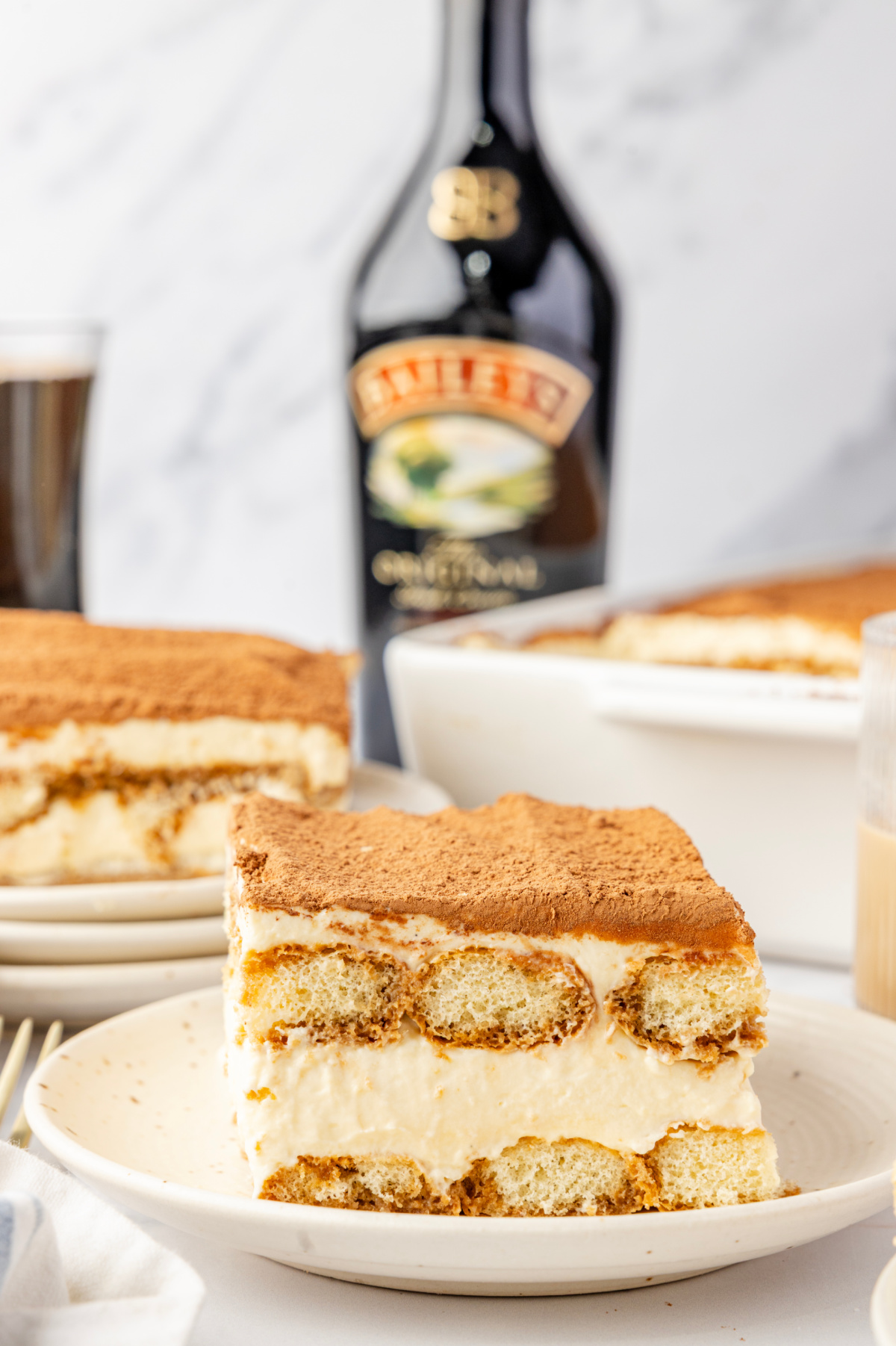 Plated slice of tiramisu with visible layers of espresso-soaked ladyfingers and mascarpone cream, with a bottle of Baileys Irish cream in the background.