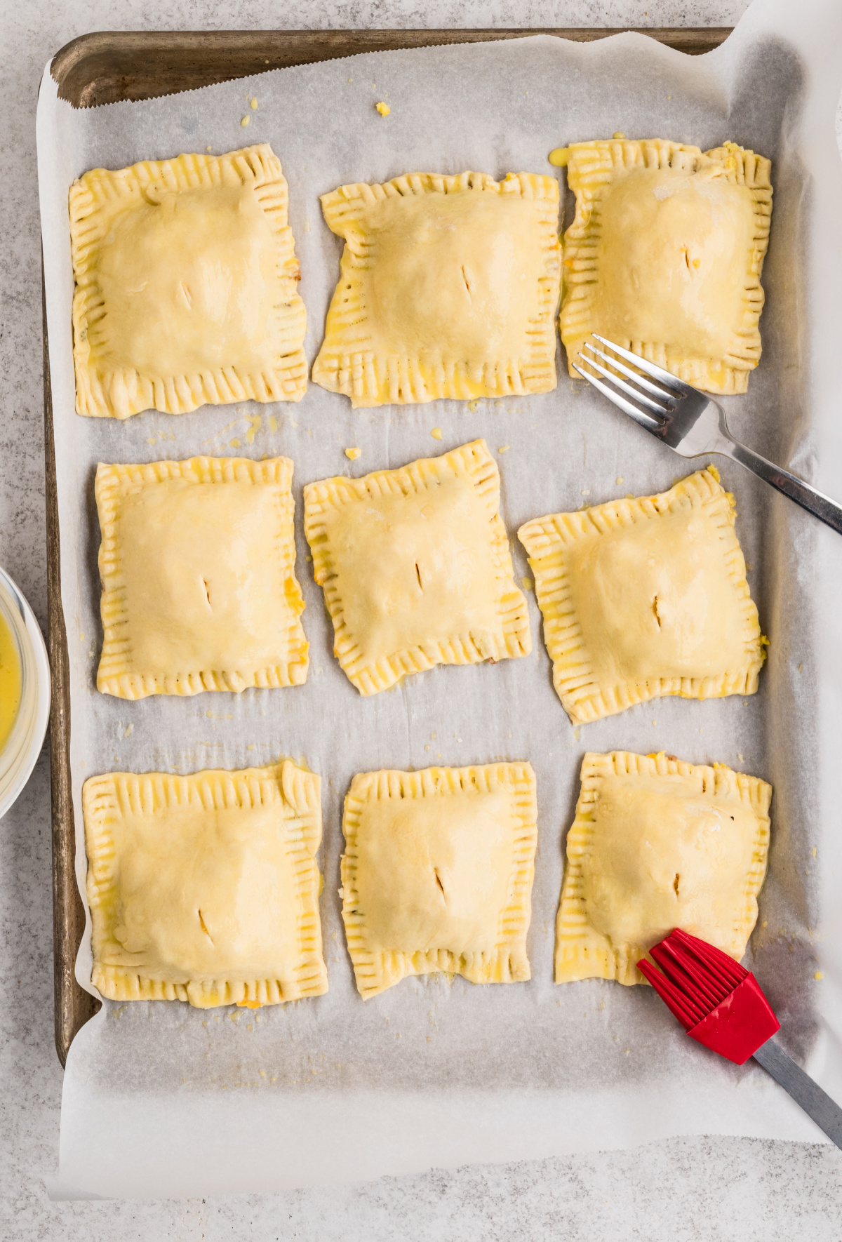 These unbaked puff pastry breakfast pastries are sealed, vented, and brushed with egg wash before ba Unbaked puff pastry breakfast pastries sealed with fork-crimped edges and brushed with egg wash on a parchment-lined baking sheet.