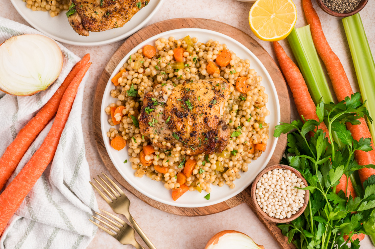 A plated serving of chicken thighs over pearl couscous with vegetables, garnished with parsley and photographed with fresh ingredients nearby. Plated chicken thighs served over pearl couscous with carrots, celery, and herbs, shown with fresh vegetables on the side.