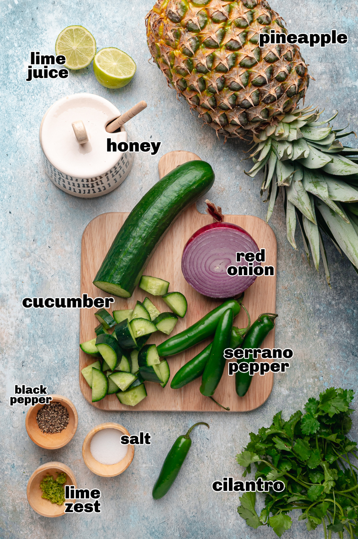 The ingredients for pineapple cucumber salad are laid out before preparation. Ingredients for pineapple cucumber salad including fresh pineapple, cucumber, red onion, serrano peppers, lime juice, lime zest, honey, salt, black pepper, and cilantro.