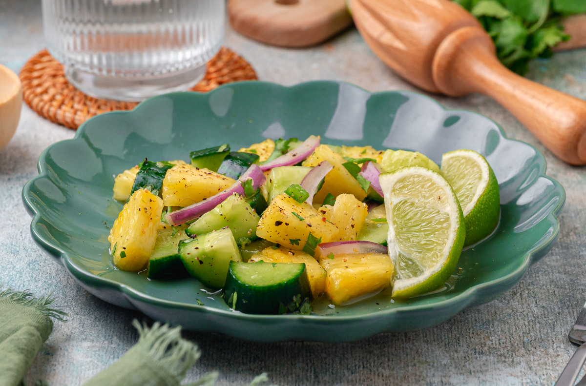 The pineapple and cucumber salad is served on a green plate with lime wedges. Pineapple and cucumber salad with lime wedges, red onion, and cilantro served on a green ceramic plate.