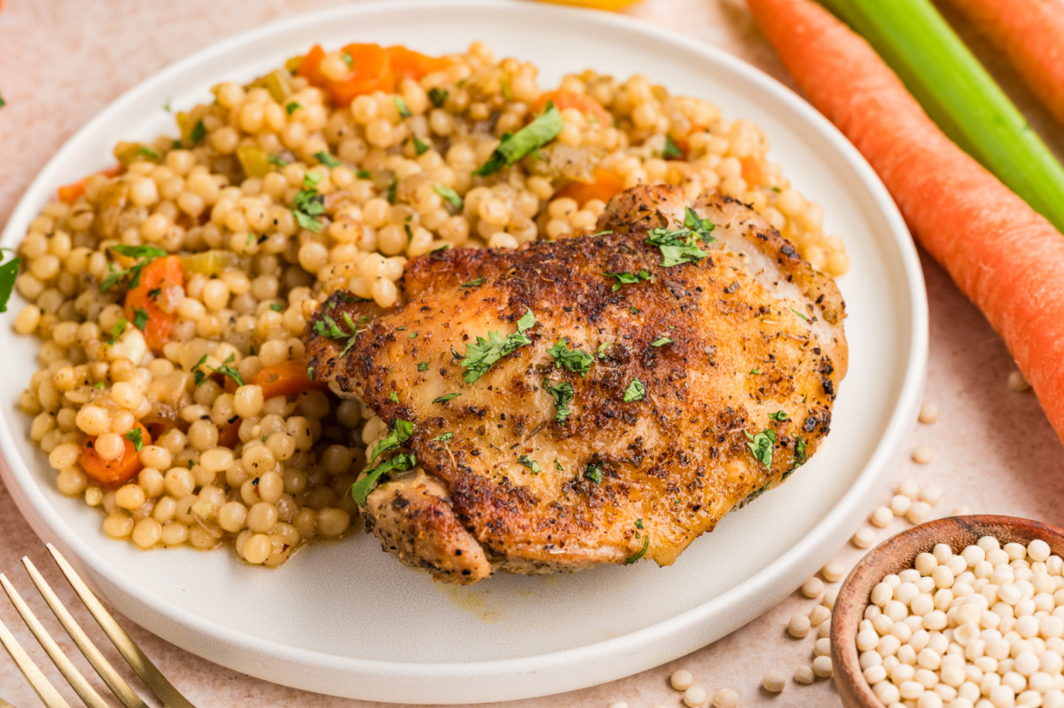 A close-up of a crispy baked chicken thigh paired with pearl couscous and tender vegetables, finished with fresh herbs. Crispy baked chicken thigh served with pearl couscous and vegetables on a plate.
