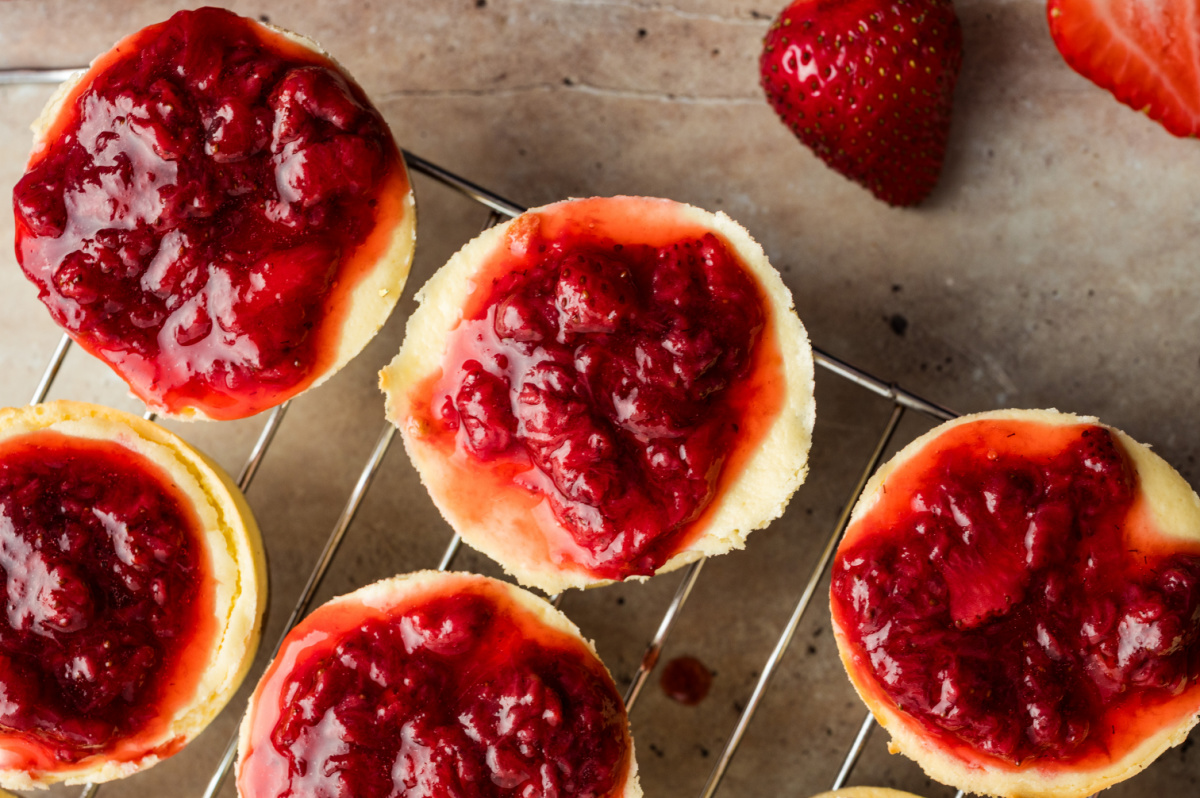 Strawberry mini cheesecakes cooling after baking with their strawberry topping set on top. Freshly baked strawberry mini cheesecakes cooling on a wire rack, each topped with a thick layer of cooked strawberry topping.