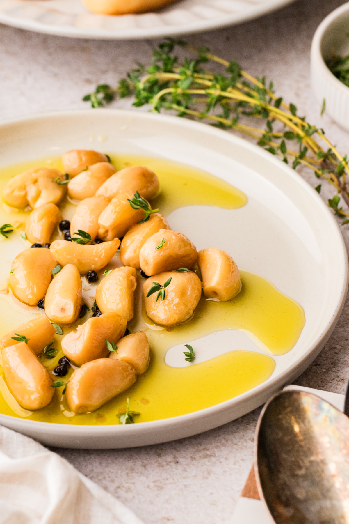 Garlic confit in olive oil with fresh thyme and whole peppercorns. Garlic confit cloves resting in olive oil with thyme and peppercorns on a white plate.