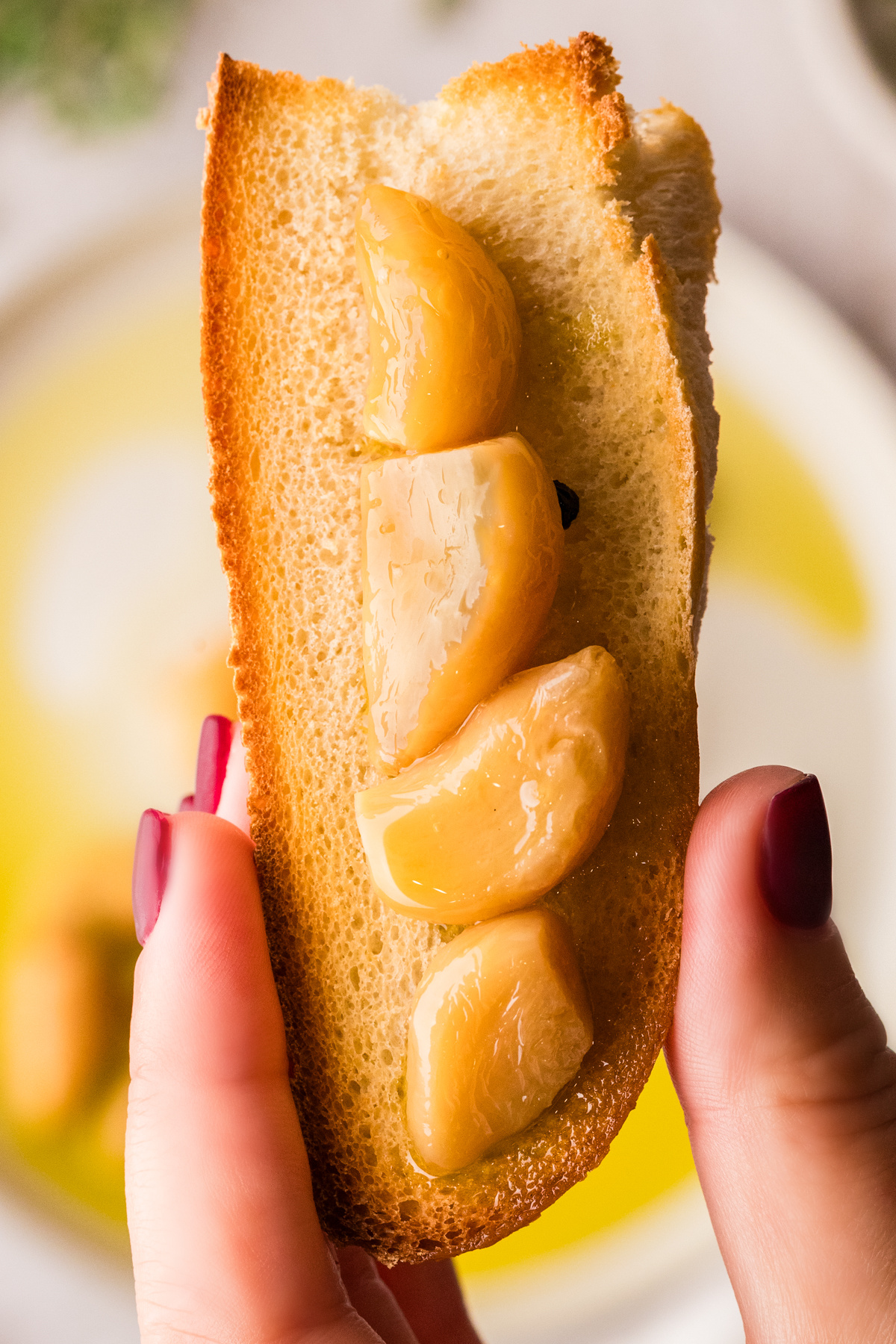 Toasted bread topped with soft oil-cooked garlic cloves. Soft garlic cloves resting on toasted bread, held upright and ready to spread.