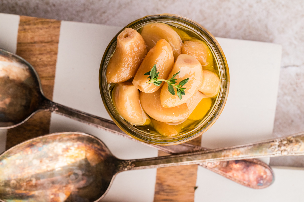 Jarred oven-cooked garlic cloves stored in olive oil. Jar of soft garlic cloves fully submerged in olive oil with thyme.