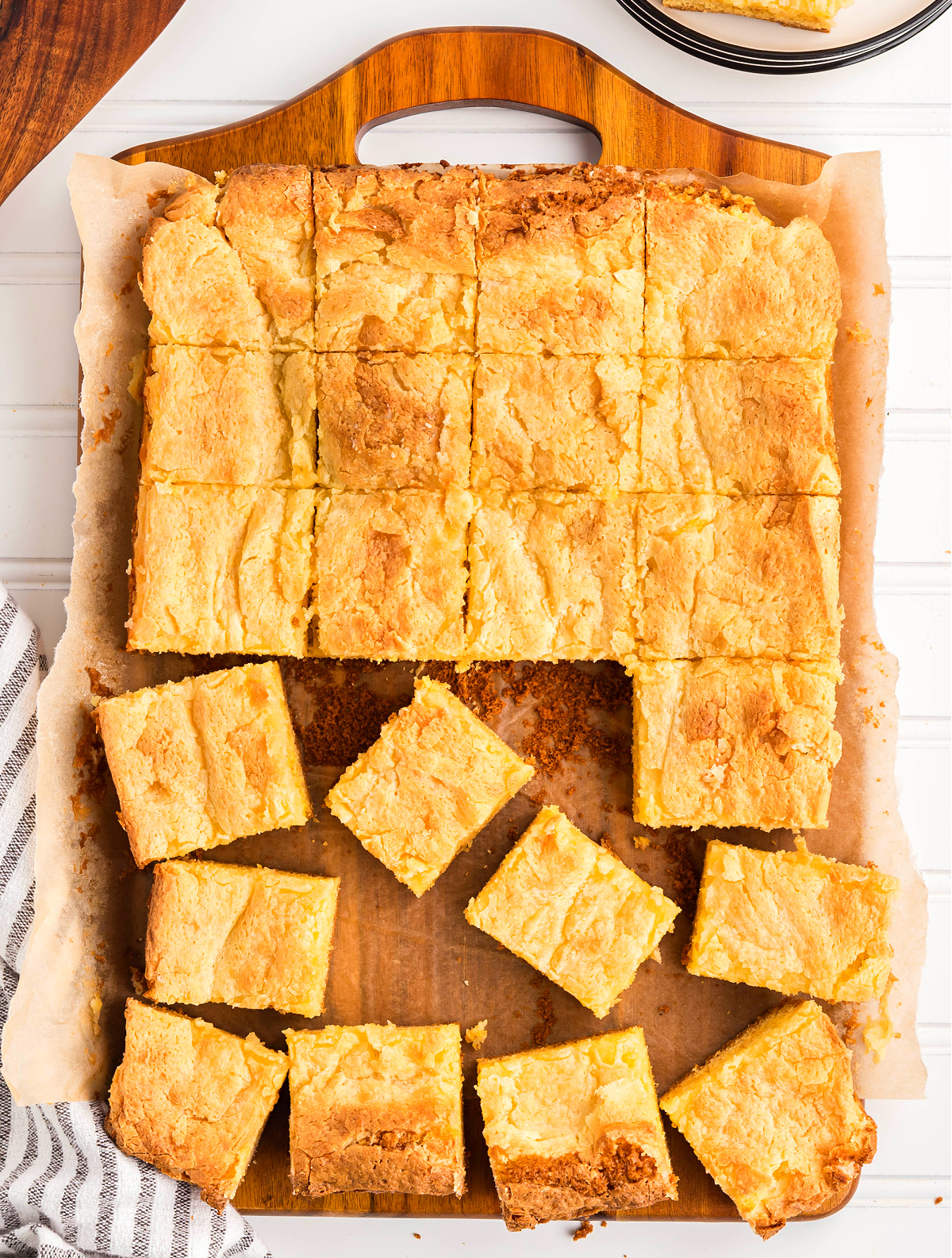 Gooey butter cake sliced into squares in a parchment-lined baking pan. Gooey butter cake baked in a parchment-lined pan and cut into soft, golden squares with a crackled top.