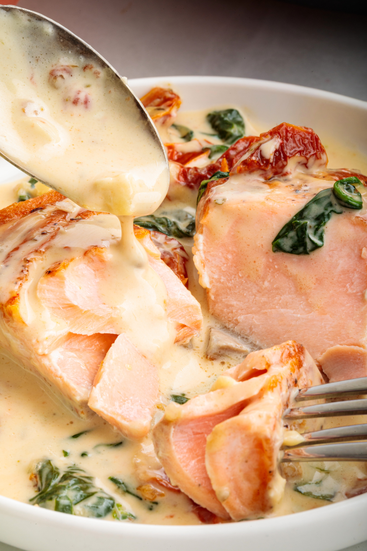 A close-up of a fork breaking into salmon fillets coated in a creamy garlic sauce with spinach and sun-dried tomatoes. Marry Me Tuscan salmon flaking apart in a creamy garlic sauce with sun-dried tomatoes and spinach.