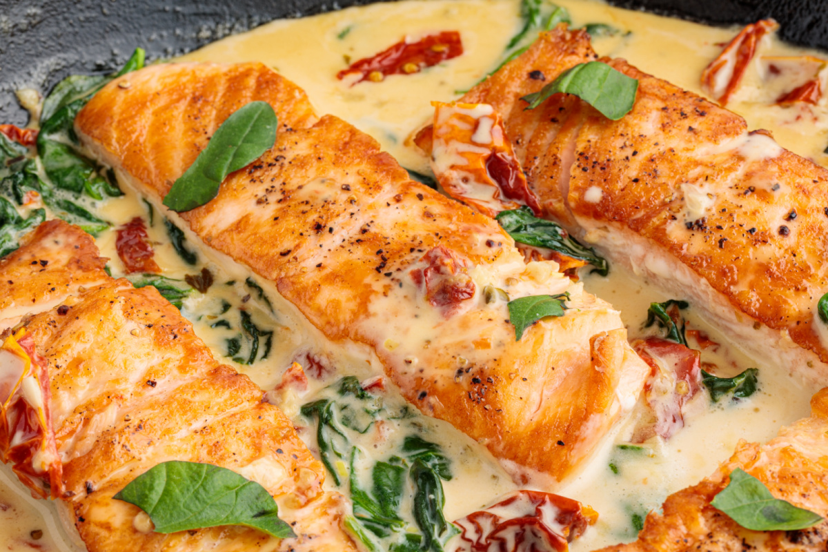 Salmon fillets simmering in a creamy garlic sauce with sun-dried tomatoes and spinach in a single skillet. One pan Tuscan salmon cooking in a skillet with a creamy garlic sauce, sun-dried tomatoes, and wilted spinach.