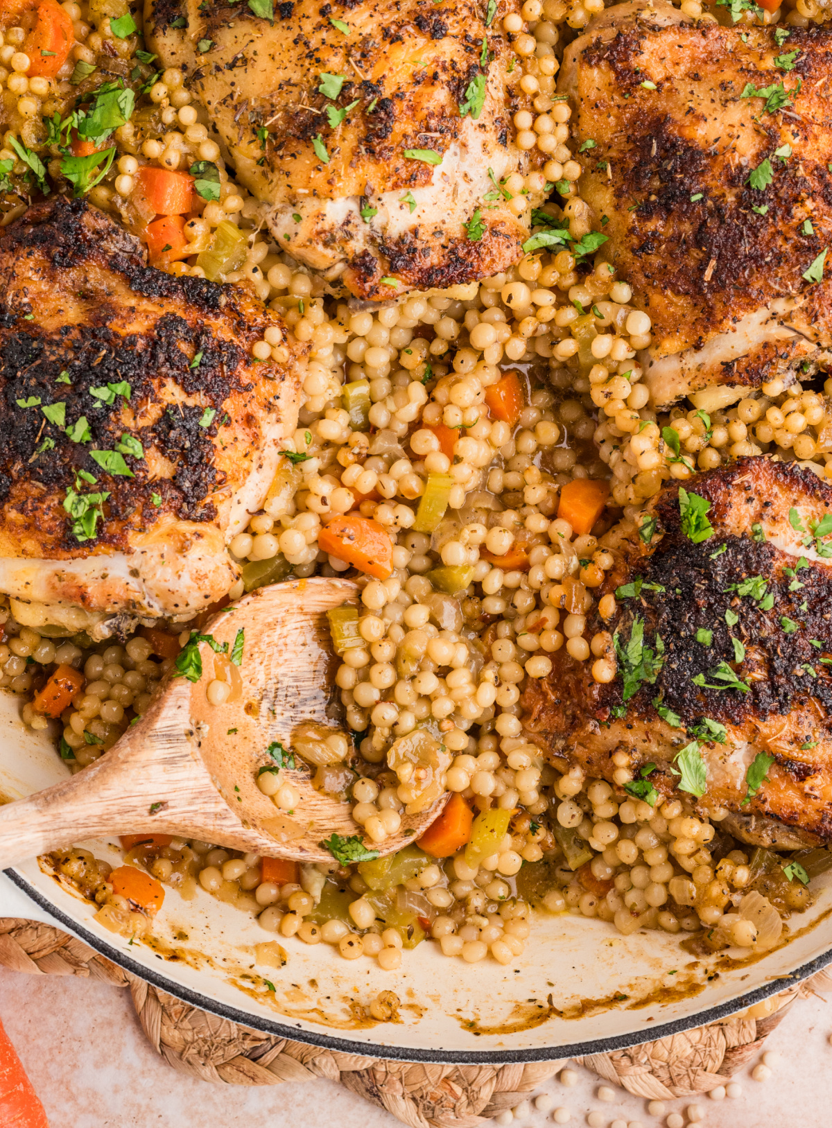 A close-up of pearl couscous scooped from the pan with crispy baked chicken thighs and vegetables cooked underneath. Spoonful of pearl couscous with vegetables served alongside crispy baked chicken thighs in a wide pan.