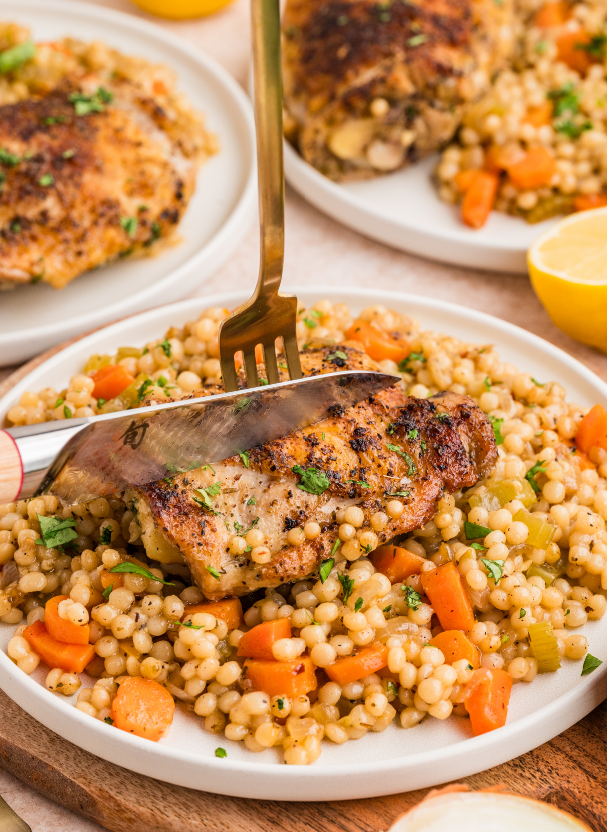 A plated serving of crispy chicken thighs over pearl couscous with vegetables, ready to eat and photographed mid-slice. Crispy chicken thighs over pearl couscous served on a plate with carrots and celery, shown being cut with a knife.
