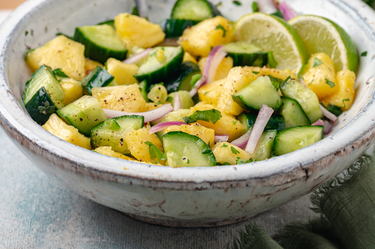 A close-up shows pineapple, cucumber, red onion, and cilantro in the salad. Close-up of pineapple and cucumber salad with red onion, cracked black pepper, cilantro, and lime slices.