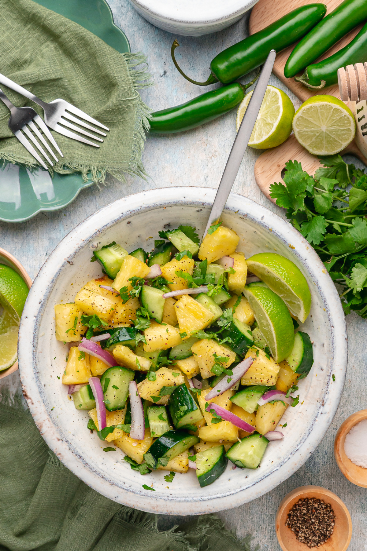 The chilled pineapple cucumber salad is finished with serrano and lime wedges. Chilled pineapple cucumber salad with fresh pineapple, cucumber, red onion, serrano pepper, cilantro, and lime wedges in a bowl.