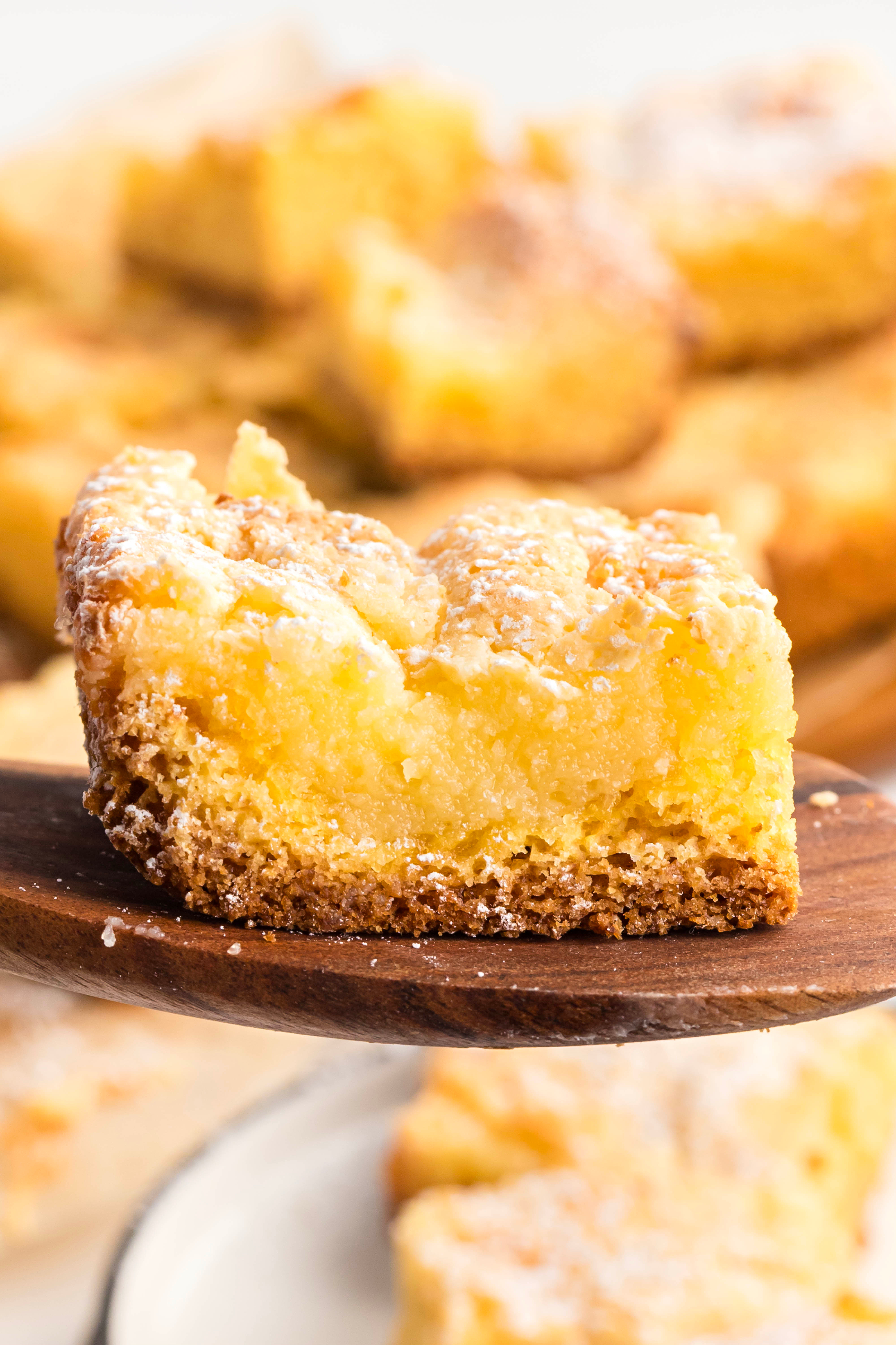 A single square of cream cheese–topped cake lifted from the pan, highlighting the gooey center and buttery base. A close-up slice of Neiman Marcus cake resting on a wooden spatula, showing the soft cream cheese layer over a dense cake base with a dusting of confectioners’ sugar.