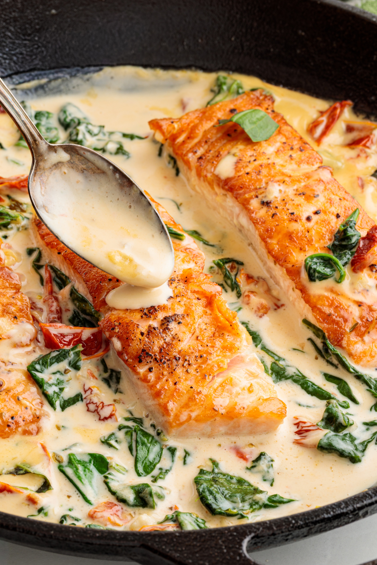 Creamy garlic sauce spooned over seared salmon fillets with spinach and sun-dried tomatoes. Salmon fillets being spooned with a creamy garlic sauce in a skillet with sun-dried tomatoes and wilted spinach.