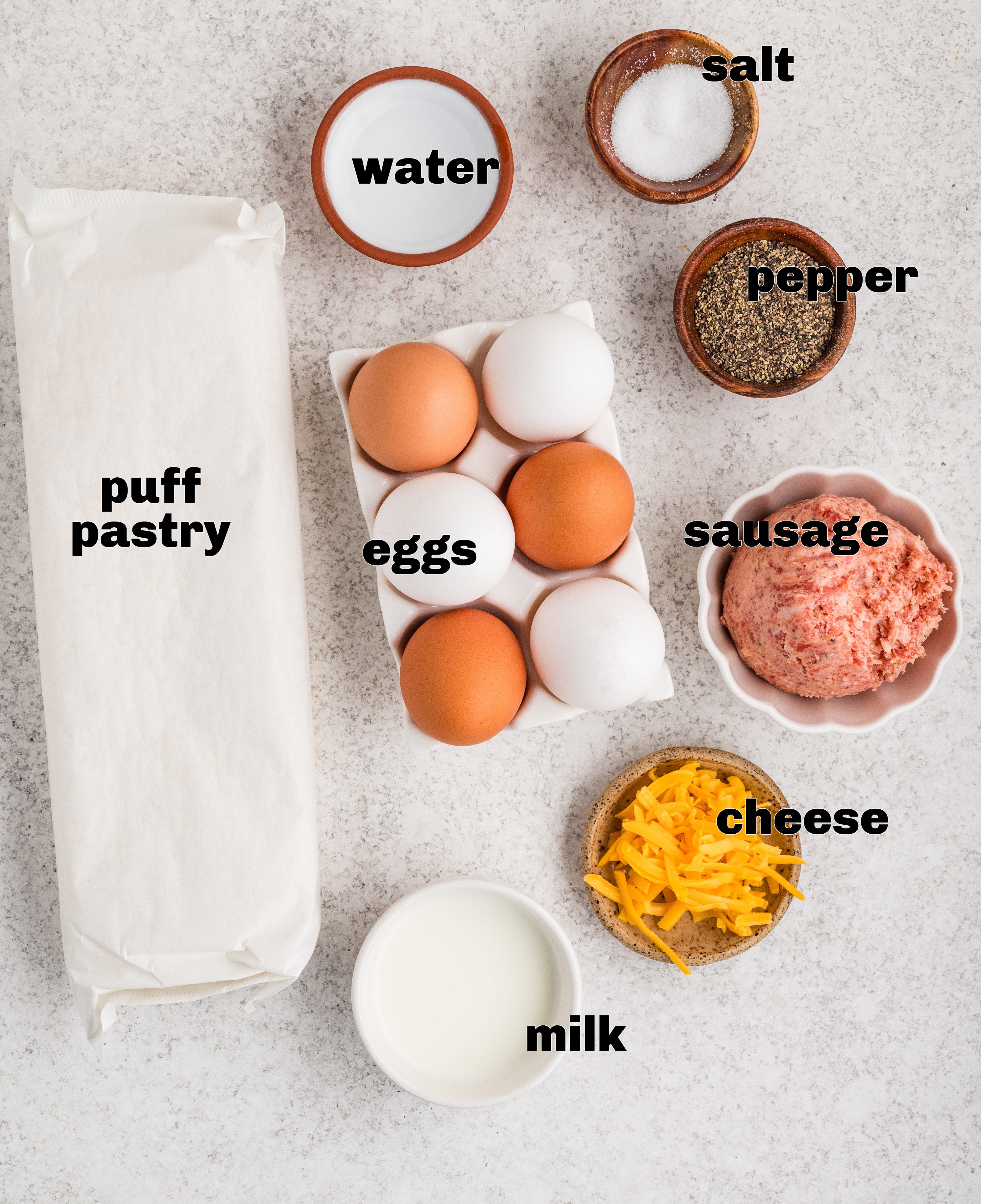 These are the simple ingredients used to make sausage, egg, and cheese breakfast pastries with puff pastry. Ingredients for sausage, egg, and cheese breakfast pastries arranged on a light surface, including puff pastry, eggs, sausage, cheddar, milk, salt, and pepper.