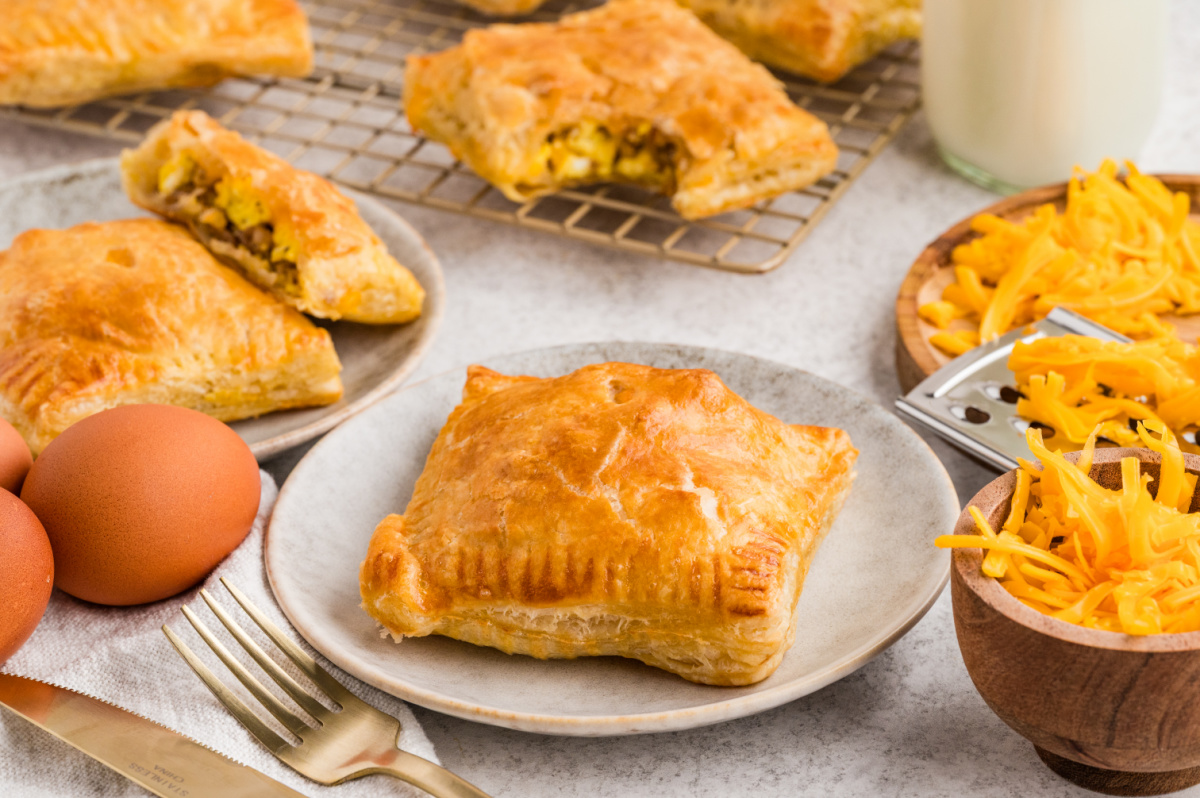 Baked puff pastry breakfast pastries filled with sausage, scrambled eggs, and cheddar served on plates with fresh eggs and cheese nearby.