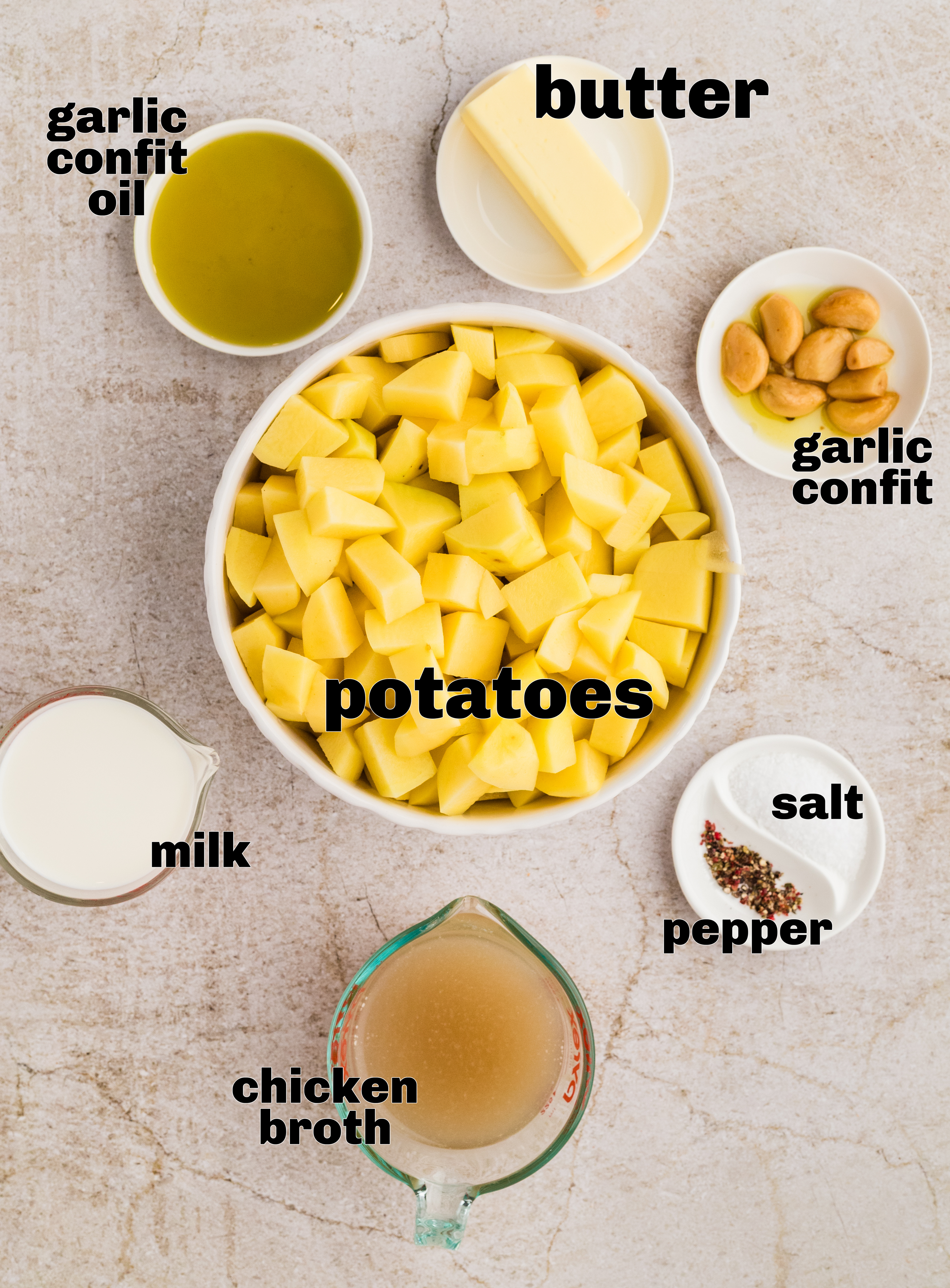 Ingredients for garlic confit mashed potatoes arranged overhead, including cubed potatoes, garlic confit cloves, garlic confit oil, butter, milk, chicken broth, salt, and pepper.