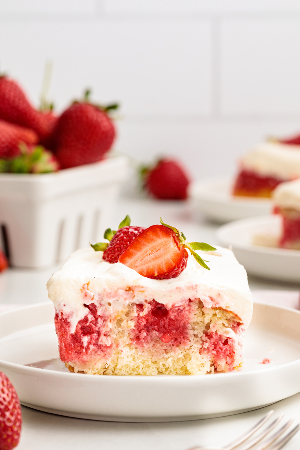This strawberry poke cake slice highlights the set strawberry filling, soft white cake, and whipped vanilla cream cheese frosting after chilling. Strawberry poke cake slice with whipped vanilla cream cheese frosting and a fresh strawberry on top, showing the soaked strawberry filling throughout the cake.