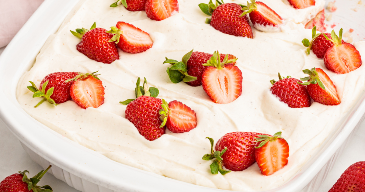 This strawberry Jell-O poke cake is finished with a smooth layer of whipped vanilla cream cheese frosting and scattered fresh strawberries. Strawberry Jell-O poke cake in a white baking dish topped with whipped vanilla cream cheese frosting and fresh strawberry halves.