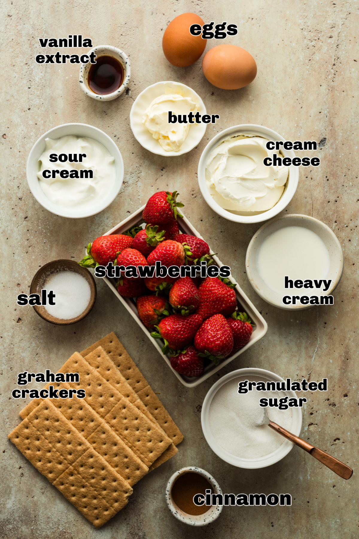 Measured ingredients laid out for making strawberry mini cheesecakes from scratch. The ingredients for strawberry mini cheesecakes arranged on a countertop, including graham crackers, cream cheese, fresh strawberries, eggs, sugar, butter, sour cream, heavy cream, vanilla, cinnamon, and salt.