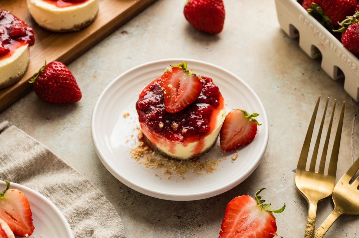 A single mini cheesecake plated with strawberry topping and a graham cracker crust. An individual mini cheesecake with a graham cracker crust and fresh strawberry topping served on a small plate with sliced strawberries nearby.