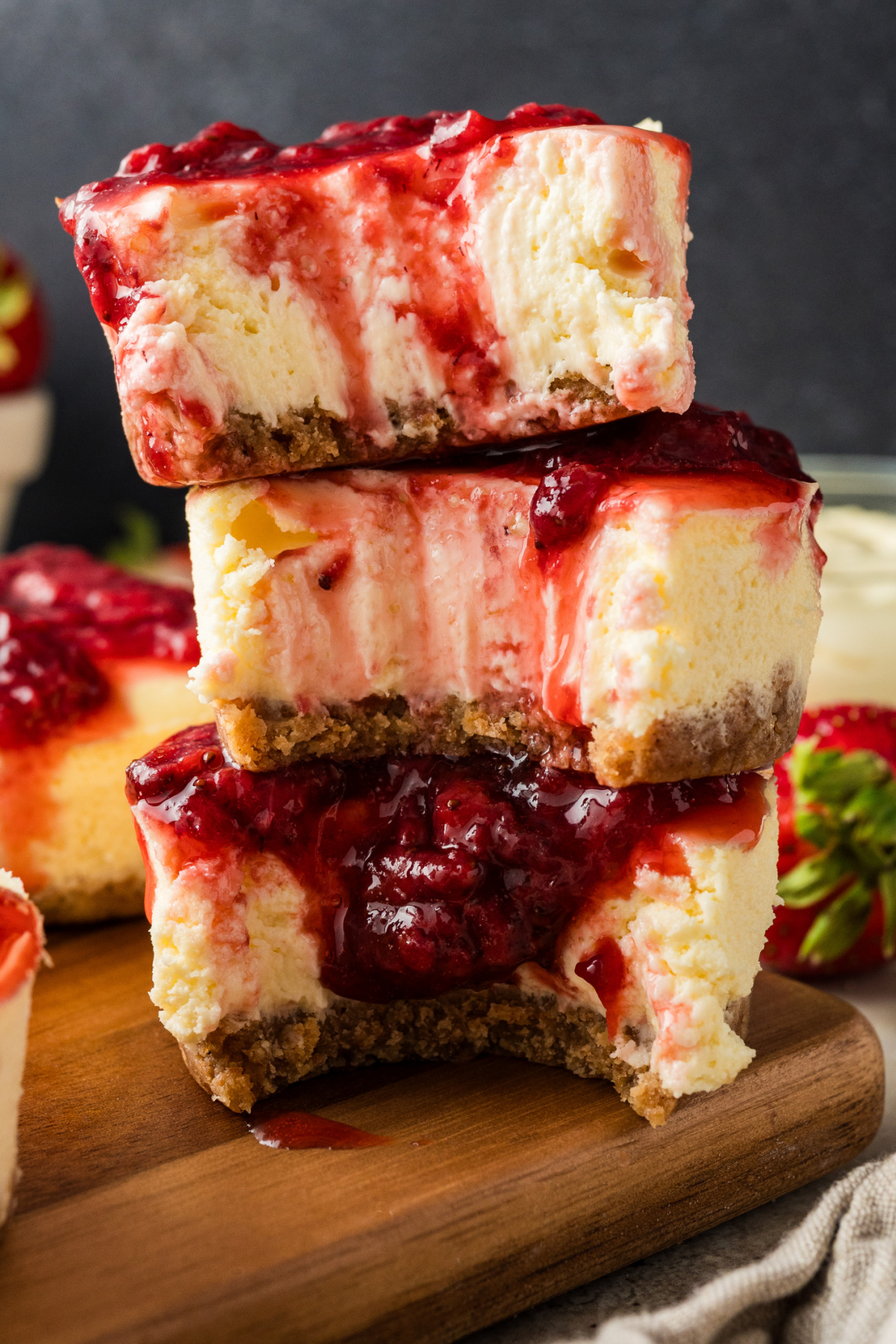 A stacked view of strawberry mini cheesecakes highlighting the creamy filling and graham cracker crust. Three strawberry mini cheesecakes stacked on a wooden board, showing the creamy cheesecake interior, graham cracker crust, and thick strawberry topping dripping down the sides.