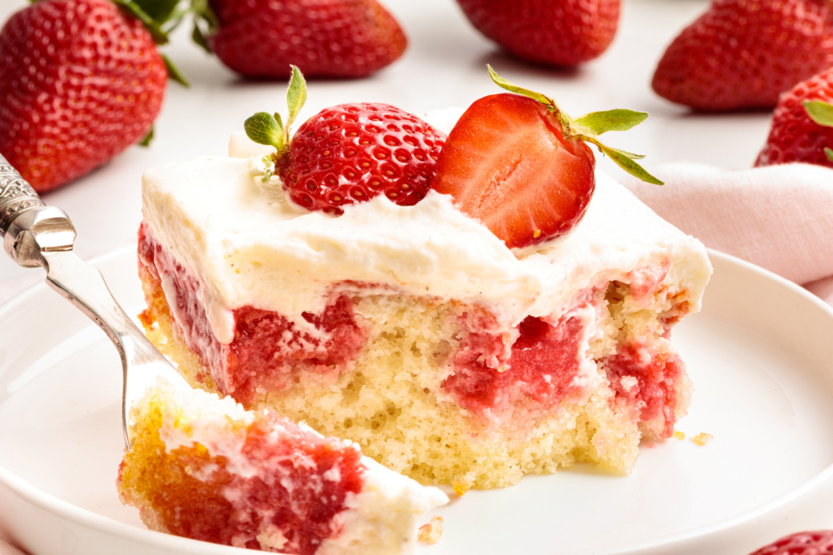 This close-up shows a bite of strawberry Jell-O poke cake with set strawberry filling and whipped vanilla cream cheese frosting. Strawberry Jell-O poke cake bite showing soaked strawberry filling, soft white cake, and whipped vanilla cream cheese frosting on a fork.