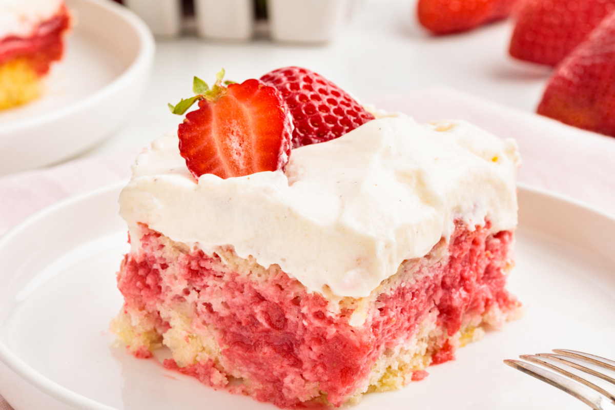 This strawberry poke cake slice shows whipped vanilla cream cheese frosting over a deeply soaked strawberry filling with visible layers. Strawberry poke cake slice with cream cheese frosting showing soaked strawberry filling layered through the cake and topped with a fresh strawberry.