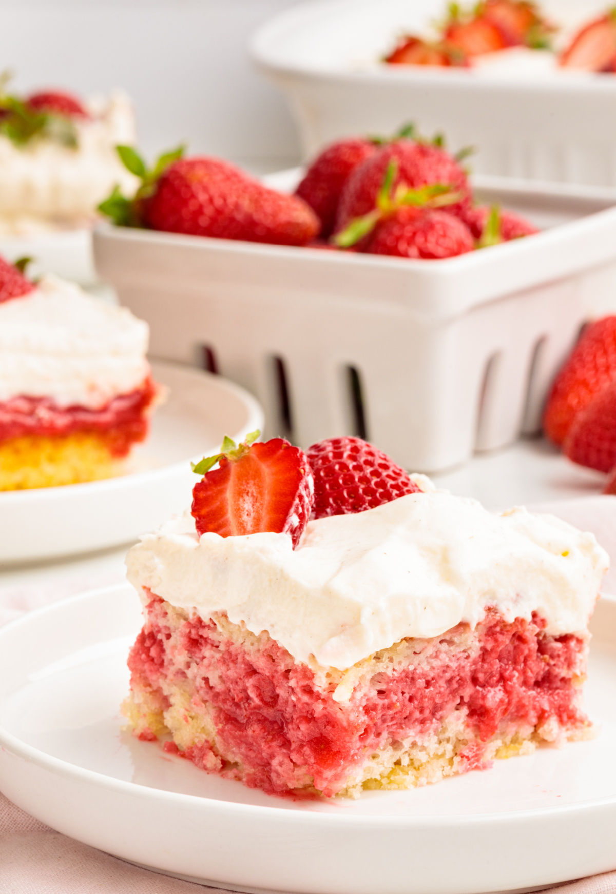 This strawberry poke cake slice shows the soaked strawberry Jell-O filling layered into the cake with whipped vanilla cream cheese frosting on top. Strawberry poke cake slice showing pink strawberry filling soaked into white cake and topped with whipped vanilla cream cheese frosting and fresh strawberries.