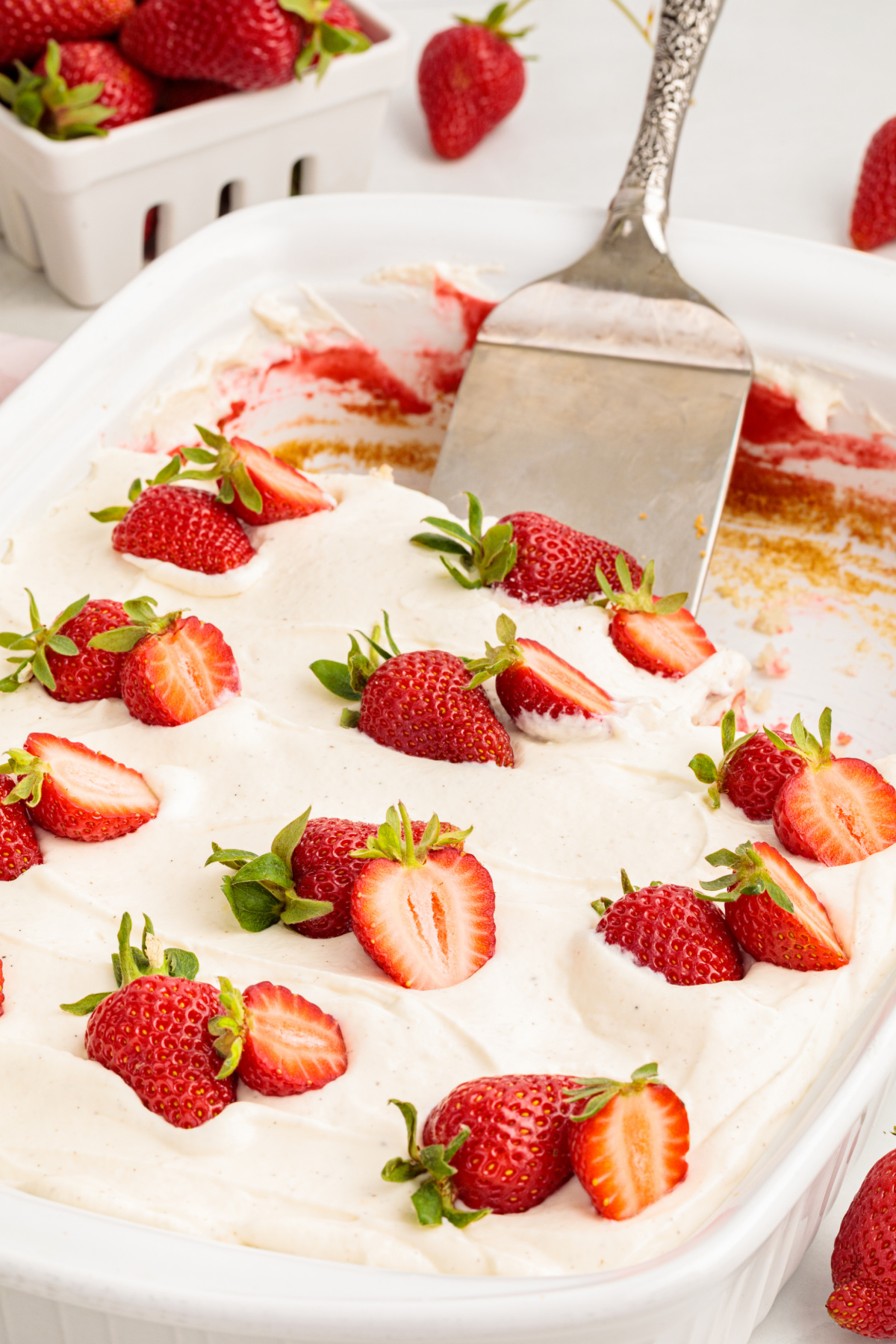 This strawberry poke cake is topped with whipped vanilla cream cheese frosting and fresh strawberries before slicing and serving. Strawberry poke cake in a baking dish with whipped vanilla cream cheese frosting spread smoothly on top and finished with fresh strawberry halves.
