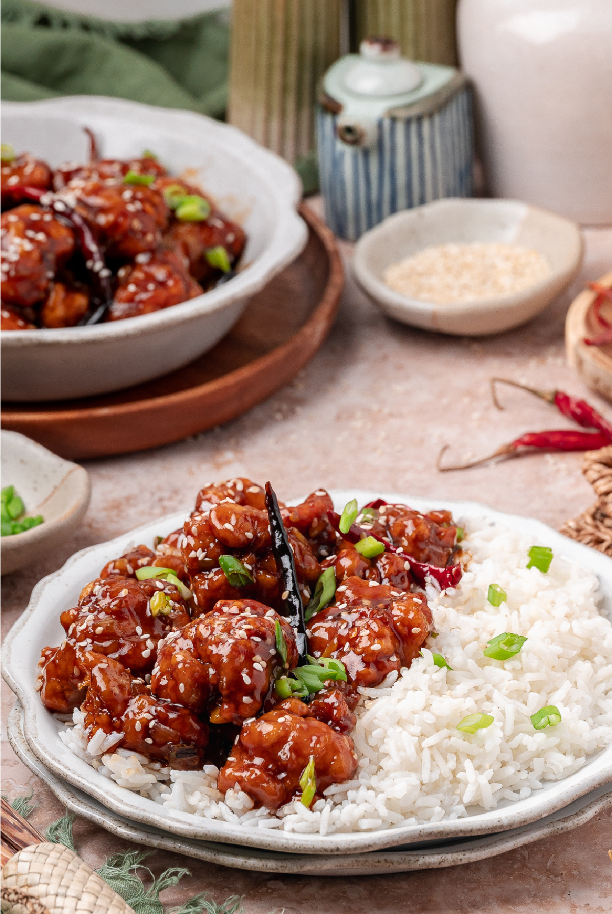 Close view of sauced fried chicken on a plate of white rice with green onions General Tso’s chicken served over white rice with scallions, sesame seeds, and dried chilies