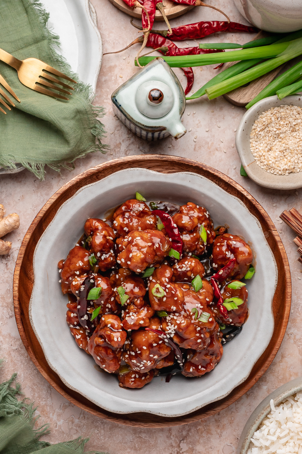 Overhead view of sauced fried chicken in a ceramic bowl with green onions and sesame garnish Bowl of crispy sweet and savory chicken topped with sesame seeds, scallions, and dried red chilies