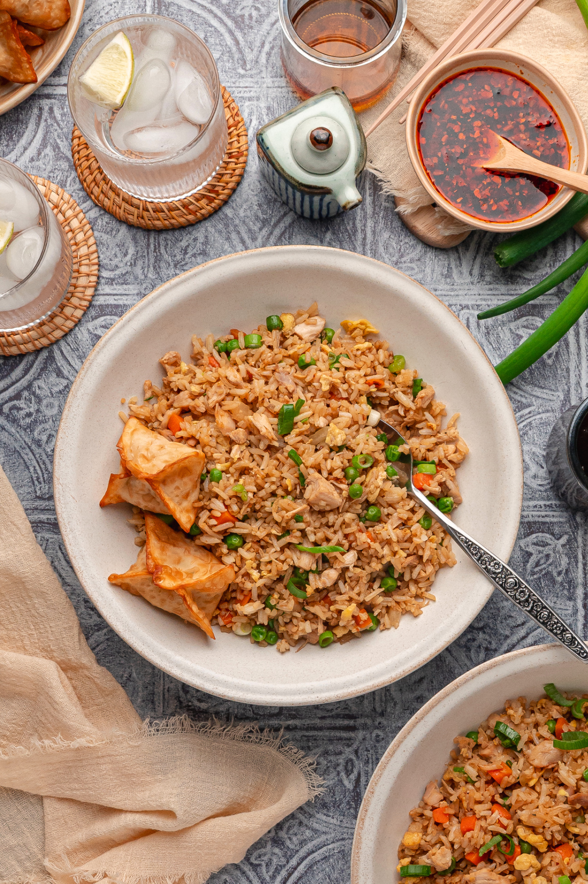 Overhead view of homemade fried rice with rotisserie chicken, scrambled eggs, peas, carrots, and sliced green onions, served in a bowl with crispy wontons and chili sauce on the table Fried rice with rotisserie chicken, eggs, peas, carrots, and green onions served with crispy wontons