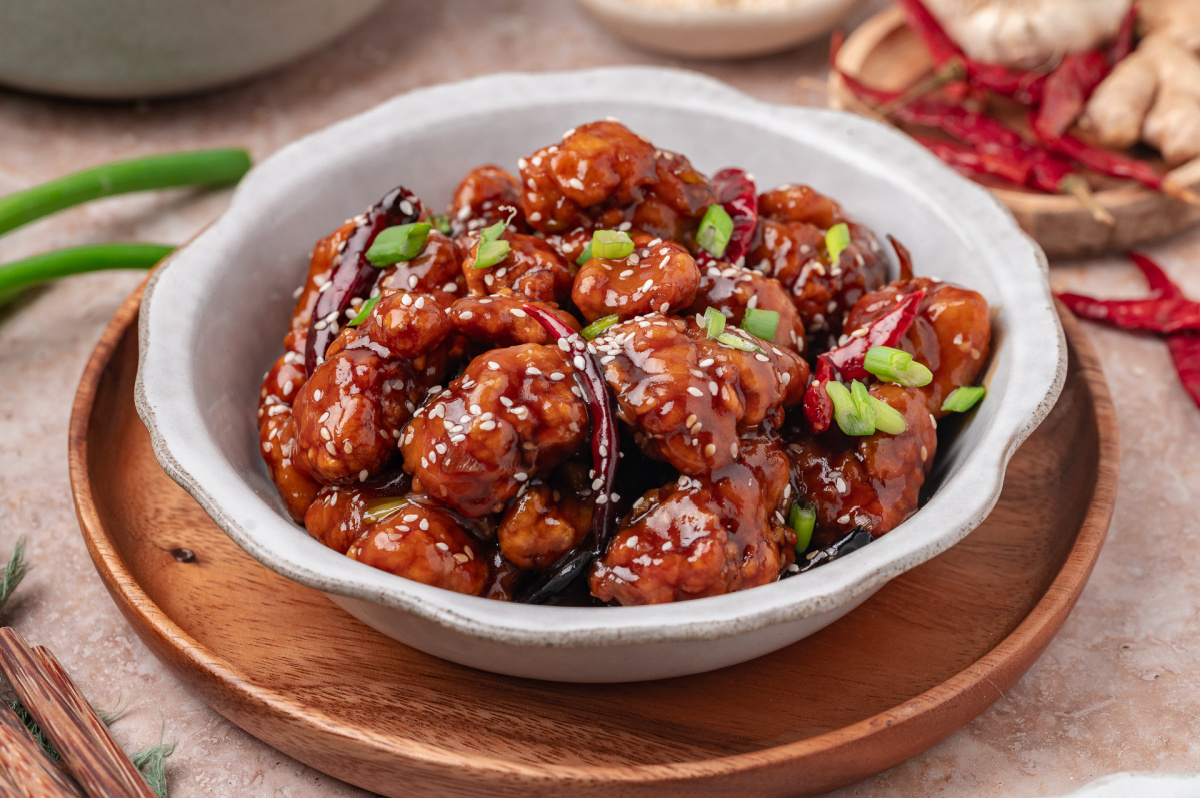 A bowl of crispy sauced chicken garnished with green onions and sesame seeds Close-up of General Tso’s chicken in a bowl with sesame seeds, scallions, and dried red chilies