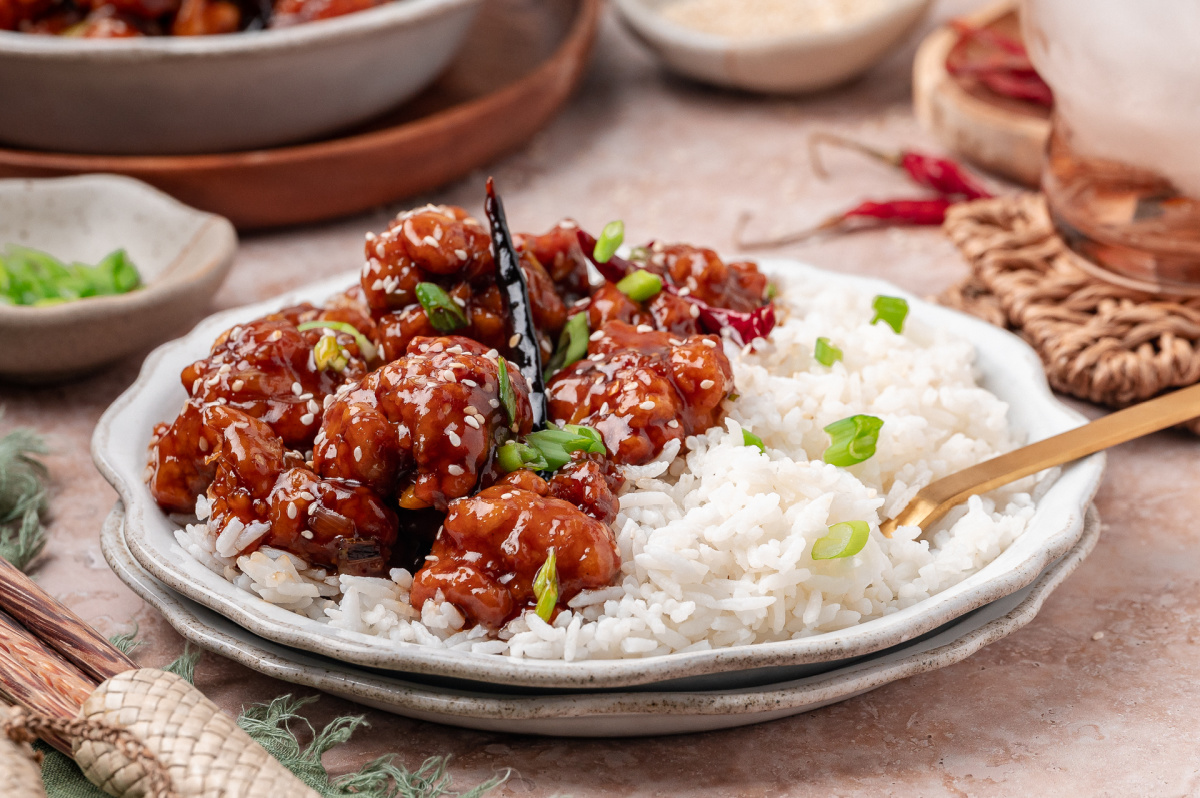 Plated crispy chicken in sweet and savory sauce alongside white rice General Tso’s chicken served over steamed white rice with scallions and sesame seeds
