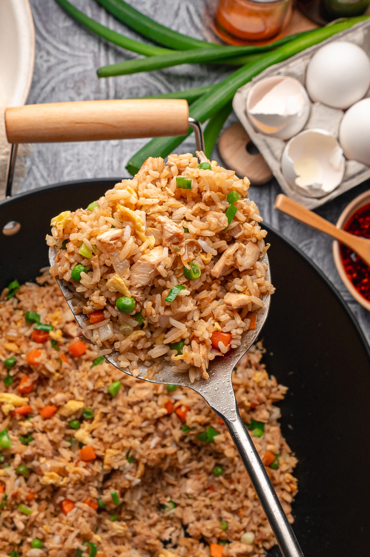Close-up of chicken fried rice being lifted from a wok with a spoon, showing separate grains of rice, rotisserie chicken, scrambled eggs, peas, carrots, and sliced green onions Chicken fried rice cooked in a wok with rotisserie chicken, eggs, peas, carrots, and green onions