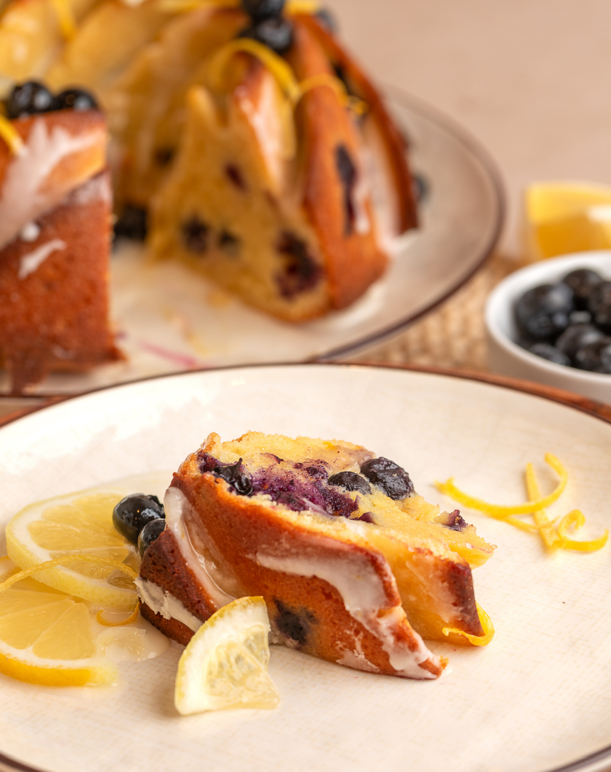 lemon blueberry bundt cake slice served with glaze and fresh fruit slice of lemon blueberry bundt cake on a plate with glaze, fresh blueberries, and lemon slices