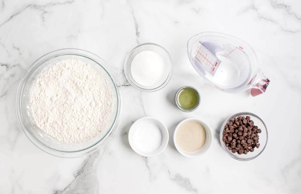 measured ingredients laid out for making chocolate chip bread ingredients for chocolate chip bread including flour, yeast, sugar, salt, oil, water, and chocolate chips on a marble surface