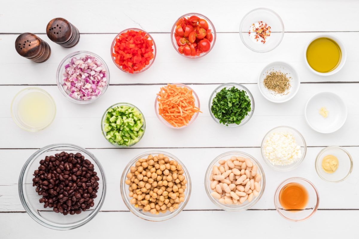 Ingredients for dense bean salad including chickpeas, black beans, cannellini beans, feta, cucumber, tomatoes, carrots, and lemon Dijon dressing