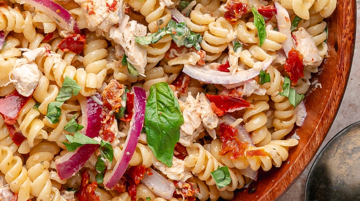 Close-up of rotini pasta salad with chicken, basil, Parmesan, and sun-dried tomatoes throughout the bowl Sun-Dried Tomato Chicken Pasta Salad with basil, rotisserie chicken, red onion, and a glossy dressing made with oil-packed sun-dried tomatoes