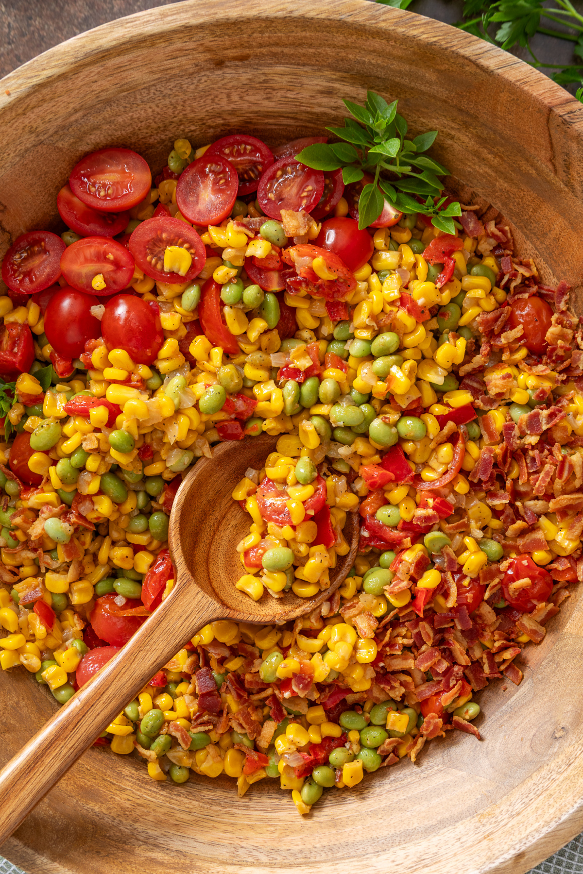 corn succatash in a wooden bowl with a spoon Overhead view of a bacon and corn side dish with edamame, tomatoes, peppers, and onions in a wooden bowl