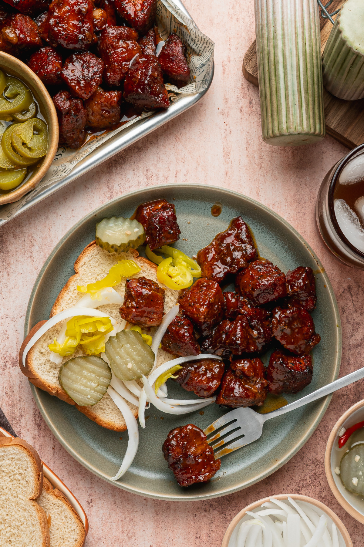 Serve oven pork belly burnt ends with bread, pickles, onions, and pepperoncini. Oven pork belly burnt ends served with white bread, pickles, onions, and pepperoncini on a dinner plate.