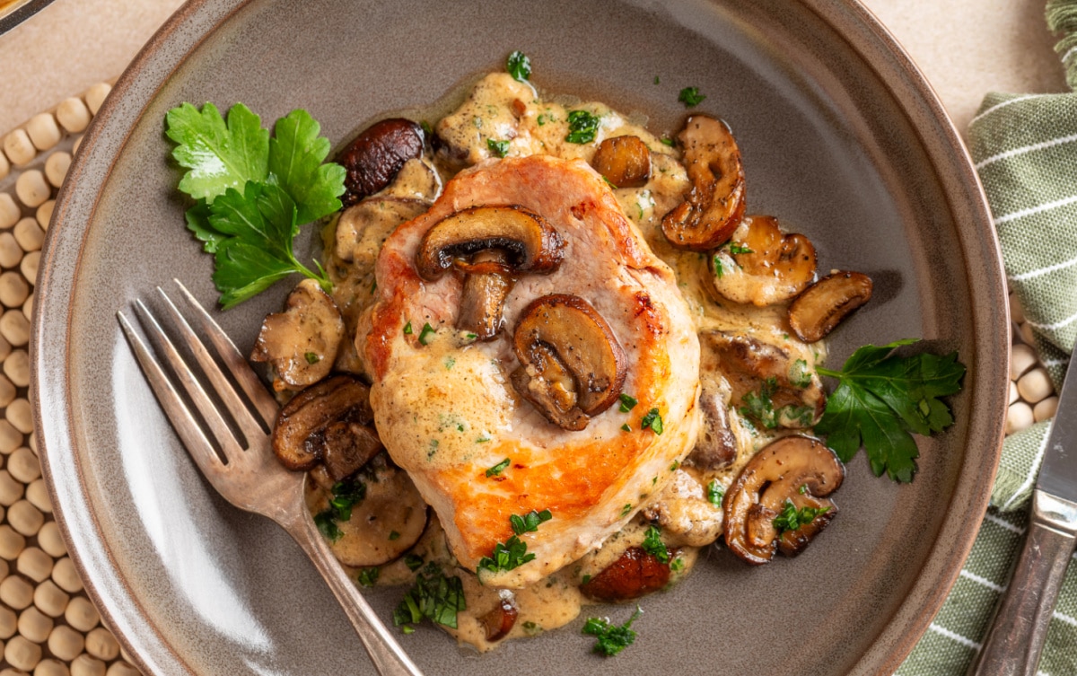 Seared boneless pork chop plated with browned mushrooms, sage cream sauce and fresh parsley. Creamy mushroom pork chop served with roasted baby portobello mushrooms and sage cream sauce