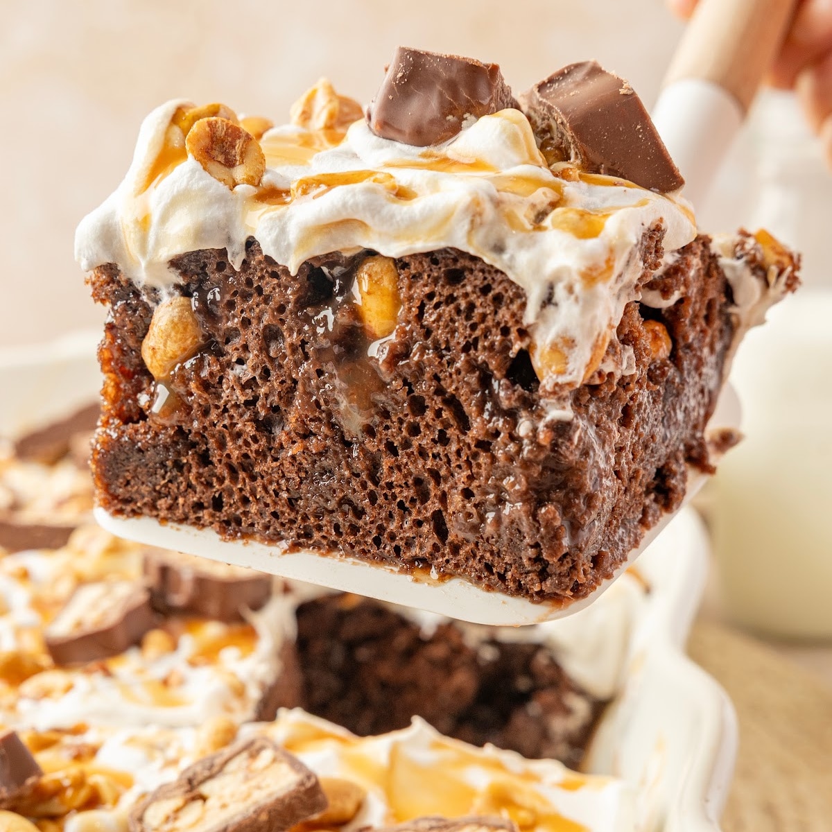 Snickers poke cake slice topped with whipped topping, caramel, salted peanuts and chopped Snickers