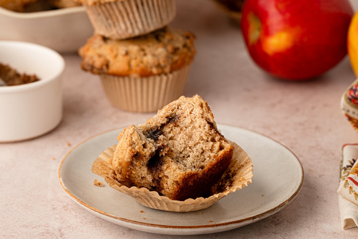 Apple cider streusel muffins made with reduced fresh cider, warm spices, brown sugar cinnamon and buttery streusel. Apple cider streusel muffin broken open on a plate with whole apples and more muffins in the background.