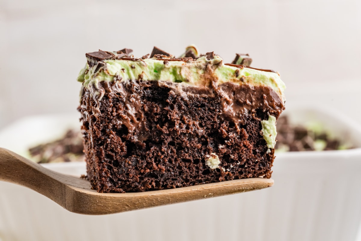 Chocolate Poke Cake with Fudge Pudding and Andes Mints Slice of chocolate poke cake on a wooden spatula showing fudge and chocolate pudding inside, topped with mint whipped cream and Andes mints.